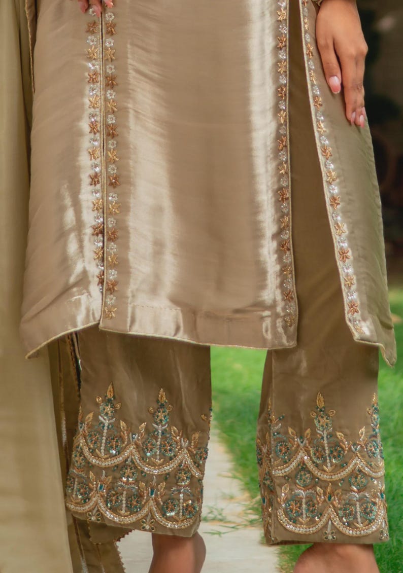 May include: A two-piece beige outfit with intricate embroidery. The top has a split front with floral embellishments. The pants feature detailed embroidery along the hemline, with gold, green, and silver accents. The fabric has a subtle sheen.