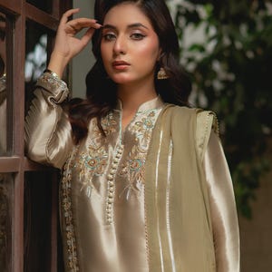 May include: A beige silk ensemble featuring a long-sleeved tunic with intricate floral embroidery in teal and gold, paired with matching trousers and a sheer scarf. The outfit has a high collar and is adorned with small, decorative buttons.