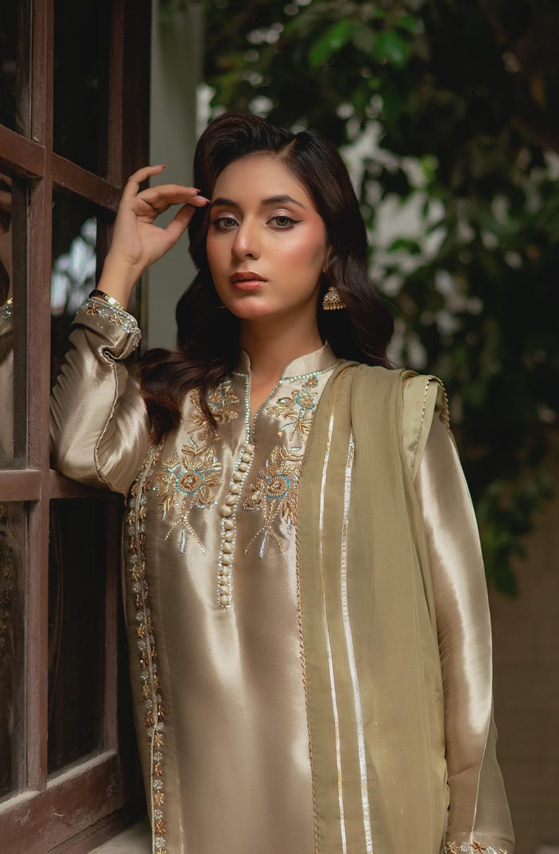 May include: A beige silk ensemble featuring a long-sleeved tunic with intricate floral embroidery in teal and gold, paired with matching trousers and a sheer scarf. The outfit has a high collar and is adorned with small, decorative buttons.
