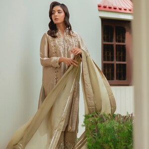 May include: A beige silk salwar kameez ensemble. The long-sleeved tunic features intricate embroidery around the neckline and cuffs. The matching trousers have detailed embroidery at the hem. A sheer, flowing dupatta completes the outfit. The model is wearing high-heeled shoes.