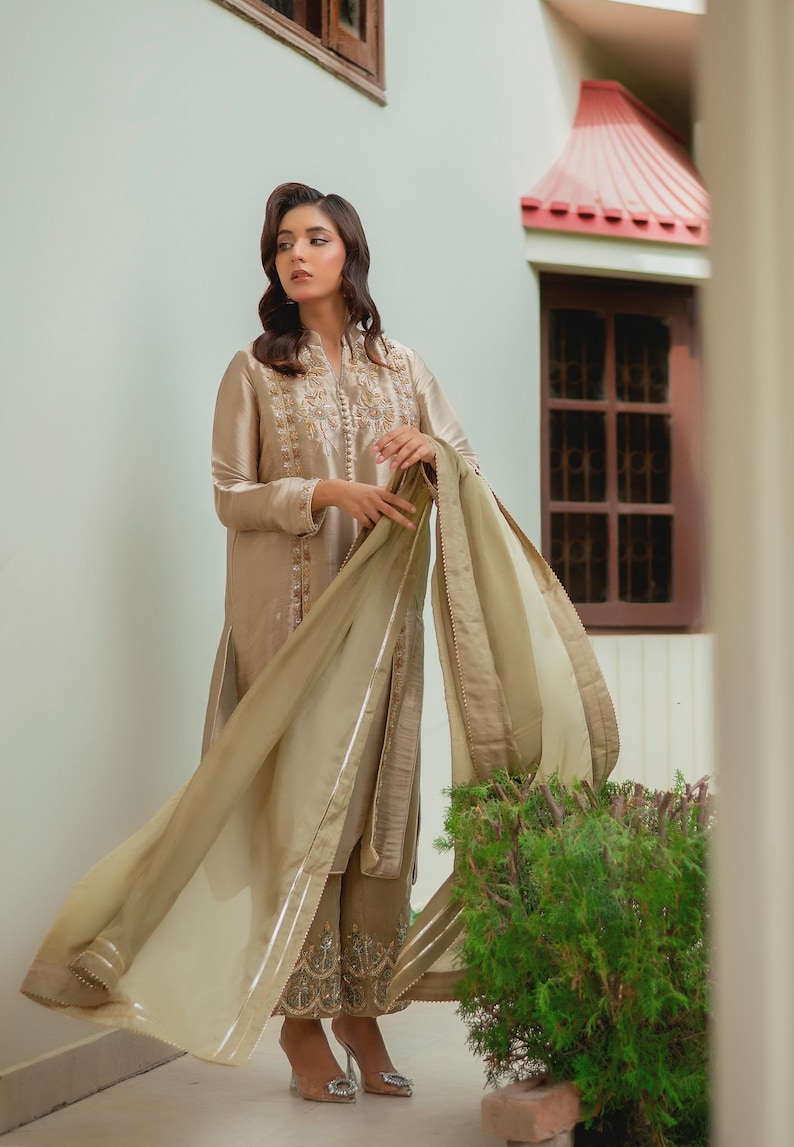 May include: A beige silk salwar kameez ensemble. The long-sleeved tunic features intricate embroidery around the neckline and cuffs. The matching trousers have detailed embroidery at the hem. A sheer, flowing dupatta completes the outfit. The model is wearing high-heeled shoes.
