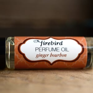 Puede incluir: Una pequeña botella de vidrio con una tapa negra y una etiqueta que dice "firebird PERFUME OIL ginger bourbon". La etiqueta es naranja con detalles marrones y una forma ovalada blanca.