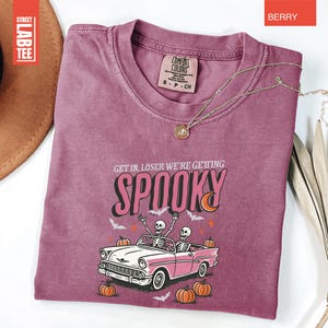 May include: Berry-colored t-shirt with the text "GET IN, LOSER WE'RE GETTING SPOOKY" and a graphic of skeletons in a pink car, pumpkins, and bats. The shirt has a crew neck and short sleeves. A gold necklace is draped across the shirt.