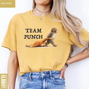 May include: Mustard-colored t-shirt with the text "TEAM PUNCH" in black. The graphic features a monkey pulling a stuffed animal. The shirt is a casual, unisex style.