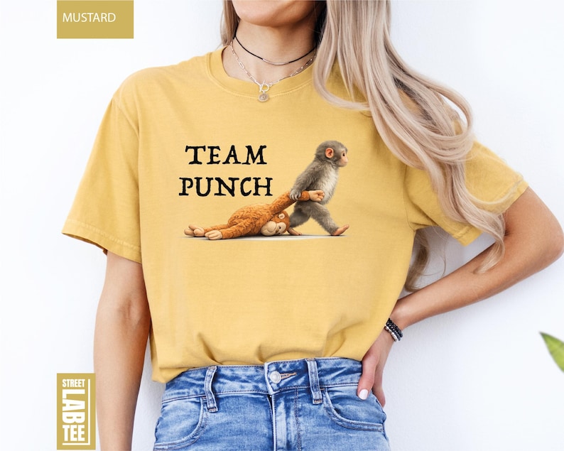 May include: Mustard-colored t-shirt with the text "TEAM PUNCH" in black. The graphic features a monkey pulling a stuffed animal. The shirt is a casual, unisex style.
