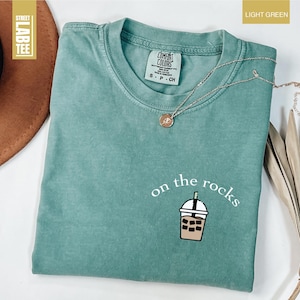 May include: Light green t-shirt with the phrase "on the rocks" and a graphic of an iced drink. The shirt has a crew neck and short sleeves. A silver necklace with a pendant is draped across the shirt.