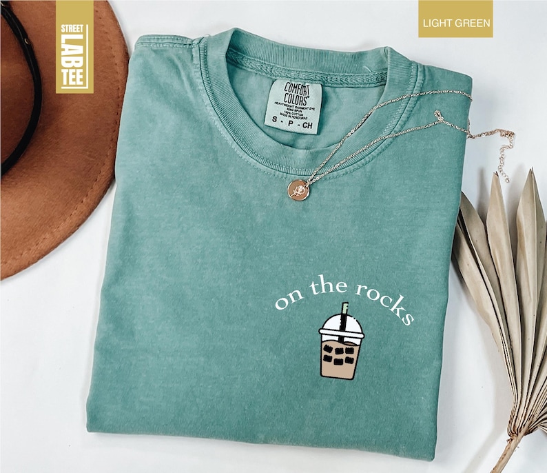 May include: Light green t-shirt with the phrase "on the rocks" and a graphic of an iced drink. The shirt has a crew neck and short sleeves. A silver necklace with a pendant is draped across the shirt.