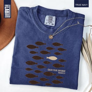 Puede incluir: Camiseta azul marino con cuello redondo. El dise&ntilde;o muestra peces marrones nadando en un grupo, con un pez color crema. El texto "CALLED TO BE DIFFERENT ROMANS 12:2" est&aacute; impreso. Una cadena dorada est&aacute; sobre la camiseta.