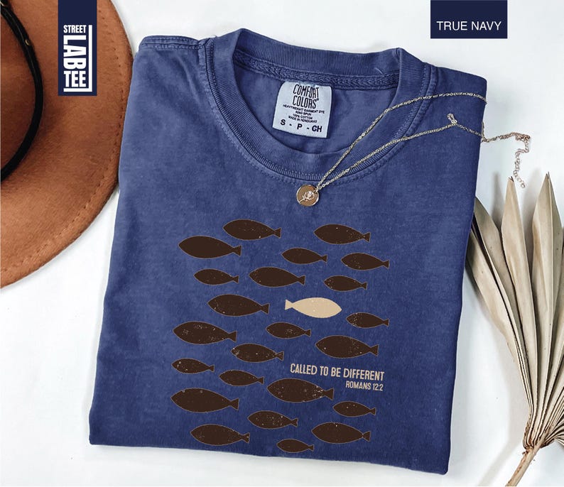 Puede incluir: Camiseta azul marino con cuello redondo. El dise&ntilde;o muestra peces marrones nadando en un grupo, con un pez color crema. El texto "CALLED TO BE DIFFERENT ROMANS 12:2" est&aacute; impreso. Una cadena dorada est&aacute; sobre la camiseta.