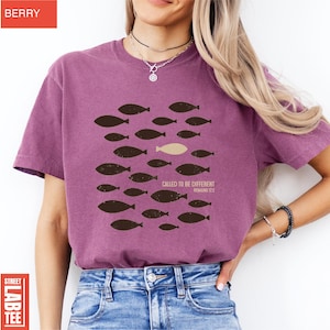 Puede incluir: Camiseta color bayas con dise&ntilde;o de peces. El dise&ntilde;o presenta un grupo de peces marr&oacute;n oscuro nadando en una direcci&oacute;n, con un pez beige nadando en la direcci&oacute;n opuesta. El texto "CALLED TO BE DIFFERENT ROMANS 12:2" est&aacute; impreso debajo.