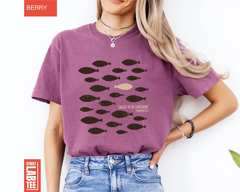 Puede incluir: Camiseta color bayas con dise&ntilde;o de peces. El dise&ntilde;o presenta un grupo de peces marr&oacute;n oscuro nadando en una direcci&oacute;n, con un pez beige nadando en la direcci&oacute;n opuesta. El texto "CALLED TO BE DIFFERENT ROMANS 12:2" est&aacute; impreso debajo.