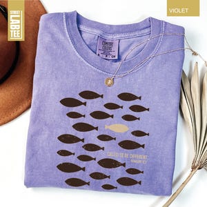 Puede incluir: Camiseta violeta con cuello redondo y un gr&aacute;fico de peces. El dise&ntilde;o presenta peces marrones nadando en grupo, con un pez beige. El texto en la camiseta dice "CALLED TO BE DIFFERENT" y "ROMANS 12:2".