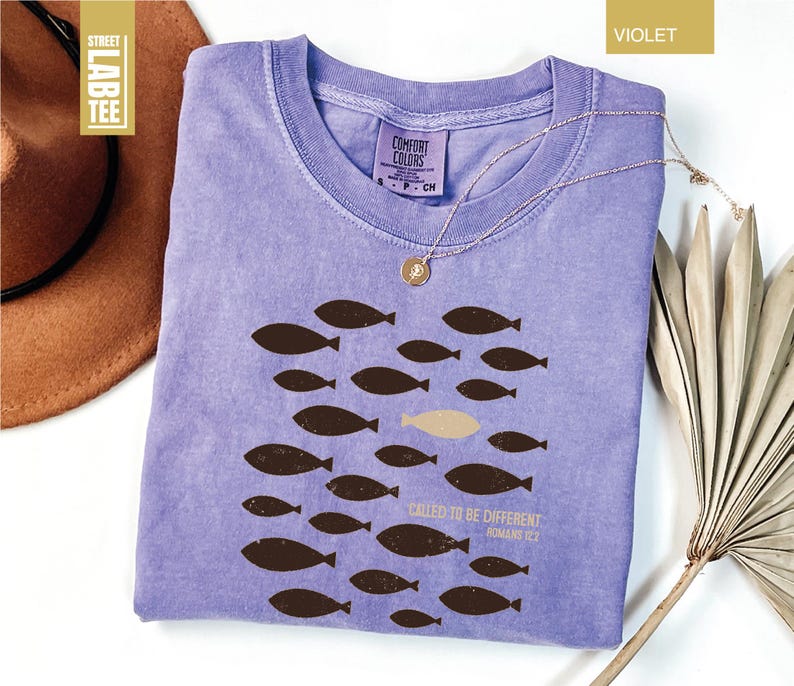 Puede incluir: Camiseta violeta con cuello redondo y un gr&aacute;fico de peces. El dise&ntilde;o presenta peces marrones nadando en grupo, con un pez beige. El texto en la camiseta dice "CALLED TO BE DIFFERENT" y "ROMANS 12:2".