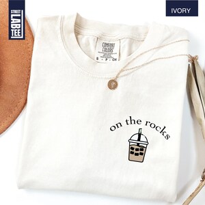 May include: Ivory-colored t-shirt with a crew neck. The shirt features the phrase "on the rocks" and a graphic of a beverage. A gold necklace with a pendant is draped on the shirt. The shirt has a tag that says "Comfort Colors".