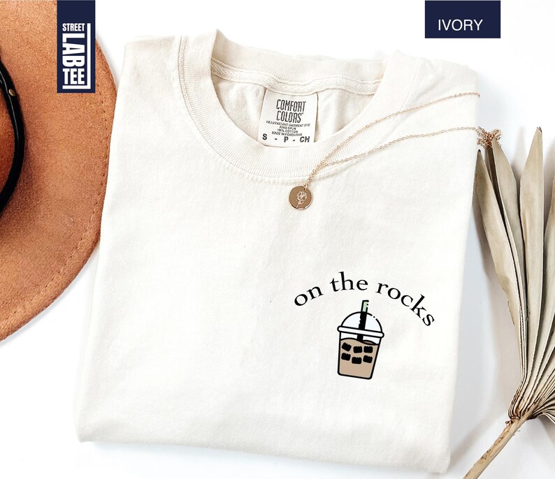 May include: Ivory-colored t-shirt with a crew neck. The shirt features the phrase "on the rocks" and a graphic of a beverage. A gold necklace with a pendant is draped on the shirt. The shirt has a tag that says "Comfort Colors".