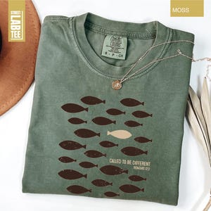 Puede incluir: Camiseta verde musgo con cuello redondo y mangas cortas. La camiseta presenta un dise&ntilde;o de peces marrones, con un pez beige, y el texto "CALLED TO BE DIFFERENT ROMANS 12:2". Un collar plateado con un colgante est&aacute; sobre la camiseta.