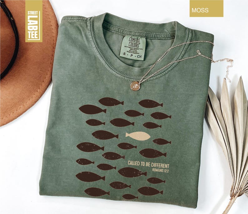 Puede incluir: Camiseta verde musgo con cuello redondo y mangas cortas. La camiseta presenta un dise&ntilde;o de peces marrones, con un pez beige, y el texto "CALLED TO BE DIFFERENT ROMANS 12:2". Un collar plateado con un colgante est&aacute; sobre la camiseta.
