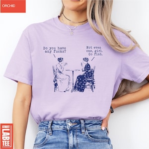May include: A light purple t-shirt with the text "Do you have any fucks?" and "Not even one, girl. Go fish." printed on it. The graphic depicts two women seated at a table. The shirt has the word "ORCHID" at the top and "STREET LAB TEE" at the bottom.