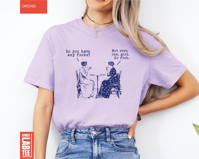 May include: A light purple t-shirt with the text "Do you have any fucks?" and "Not even one, girl. Go fish." printed on it. The graphic depicts two women seated at a table. The shirt has the word "ORCHID" at the top and "STREET LAB TEE" at the bottom.
