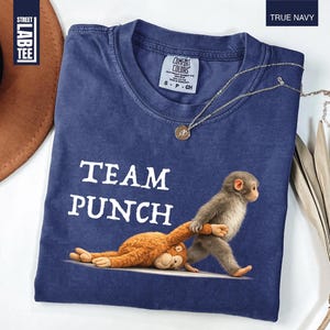 May include: True navy blue t-shirt with the text "TEAM PUNCH" and a graphic of a monkey pulling another monkey. The shirt has a crew neck and short sleeves. A gold necklace with a pendant is on the shirt.