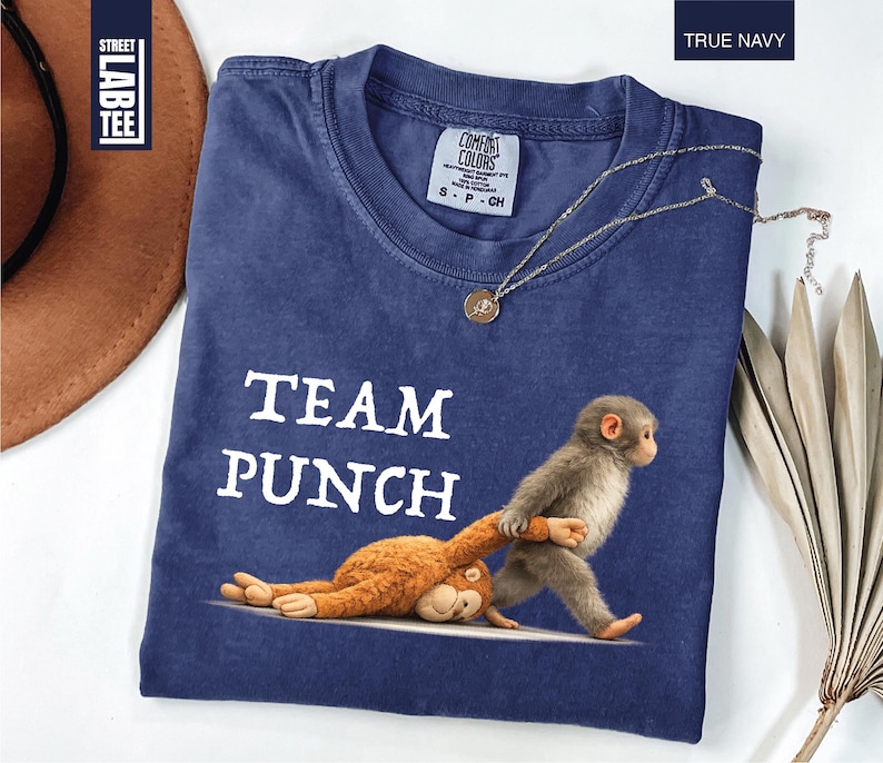 May include: True navy blue t-shirt with the text "TEAM PUNCH" and a graphic of a monkey pulling another monkey. The shirt has a crew neck and short sleeves. A gold necklace with a pendant is on the shirt.