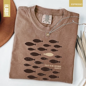 Puede incluir: Camiseta color espresso con cuello redondo y mangas cortas. La camiseta presenta un gr&aacute;fico de peces, con un pez de color m&aacute;s claro. El texto "CALLED TO BE DIFFERENT ROMANS 12:2" est&aacute; impreso debajo del gr&aacute;fico de peces. Un collar plateado est&aacute; sobre la camiseta.