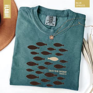 Puede incluir: Camiseta azul abeto con cuello redondo y mangas cortas. La camiseta presenta un dise&ntilde;o de peces marrones, con un pez beige, y el texto "CALLED TO BE DIFFERENT ROMANS 12:2". Un collar dorado est&aacute; drapeado sobre la camiseta.