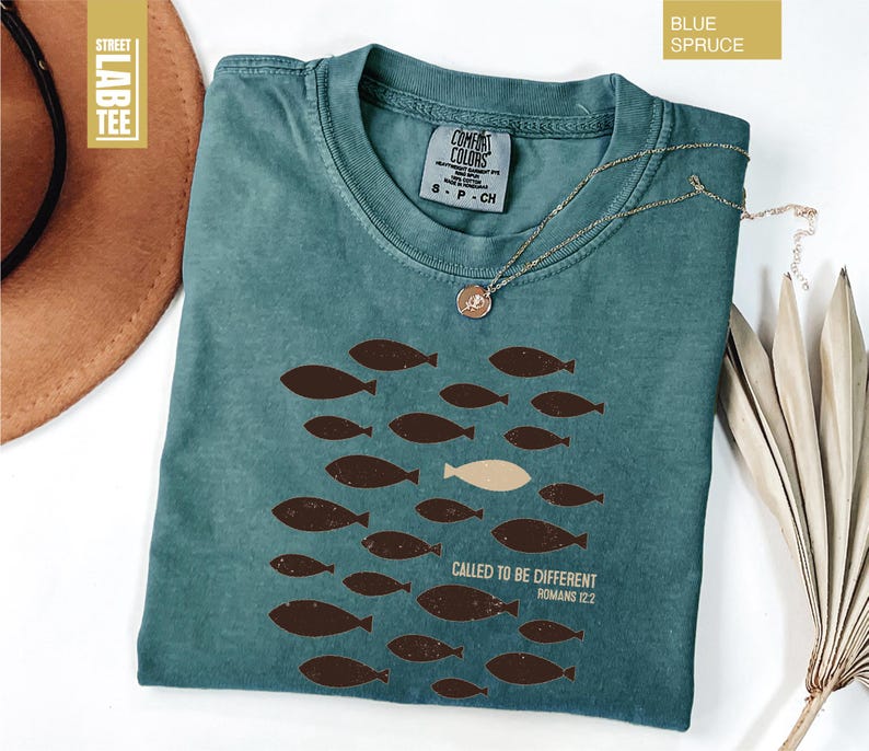 Puede incluir: Camiseta azul abeto con cuello redondo y mangas cortas. La camiseta presenta un dise&ntilde;o de peces marrones, con un pez beige, y el texto "CALLED TO BE DIFFERENT ROMANS 12:2". Un collar dorado est&aacute; drapeado sobre la camiseta.