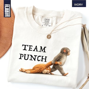 May include: Ivory-colored t-shirt with the text "TEAM PUNCH" in black. The shirt features a cartoon illustration of two monkeys. One monkey is pulling the other by the arm. A gold necklace is draped on the shirt.