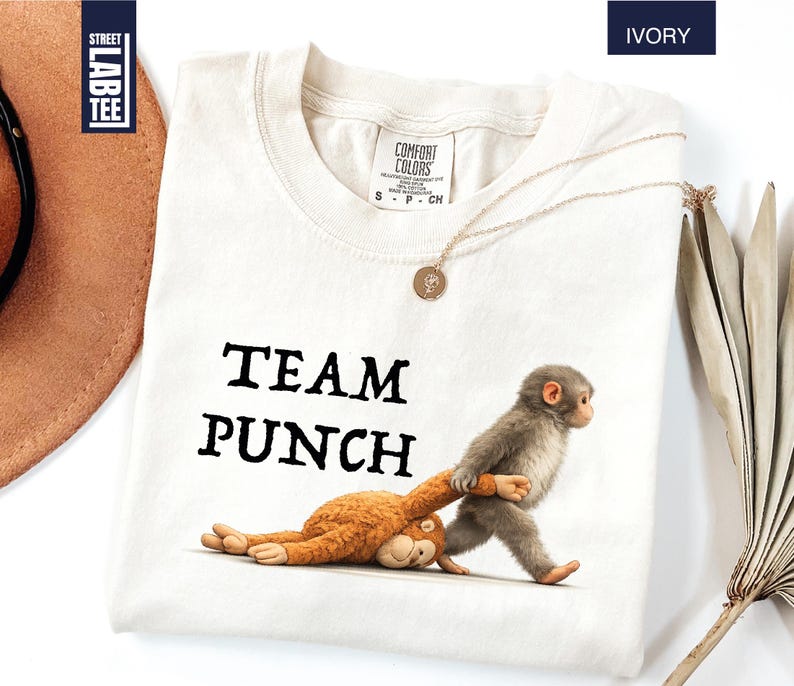 May include: Ivory-colored t-shirt with the text "TEAM PUNCH" in black. The shirt features a cartoon illustration of two monkeys. One monkey is pulling the other by the arm. A gold necklace is draped on the shirt.