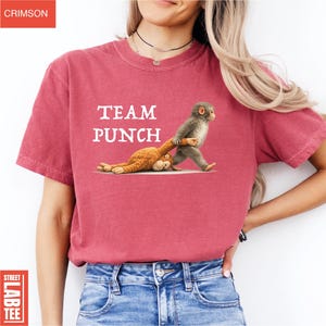 May include: Crimson red t-shirt with the text "TEAM PUNCH" in white. The shirt features a graphic of a monkey dragging a stuffed animal. The shirt is made of a soft material.