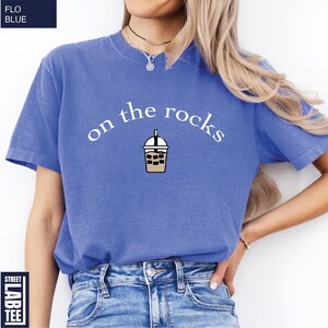 May include: A cornflower blue t-shirt featuring the phrase "on the rocks" above an iced coffee graphic. The shirt has short sleeves and is made of a soft material. The text and graphic are in white and brown.