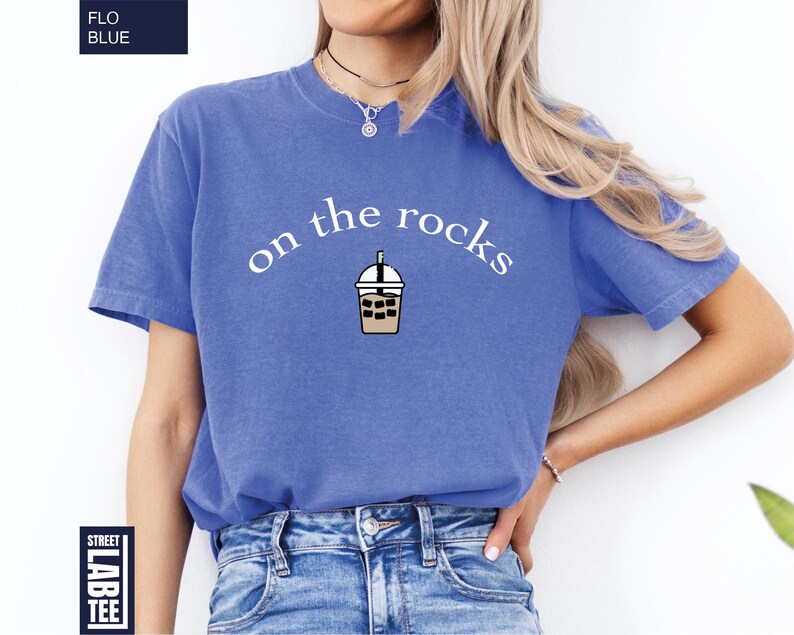 May include: A cornflower blue t-shirt featuring the phrase "on the rocks" above an iced coffee graphic. The shirt has short sleeves and is made of a soft material. The text and graphic are in white and brown.