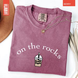 May include: Berry-coloured t-shirt featuring the words "on the rocks" and an iced beverage graphic. The shirt has a crew neck and short sleeves. A gold necklace with a pendant is draped across the shirt. The shirt is labelled "Comfort Colors".
