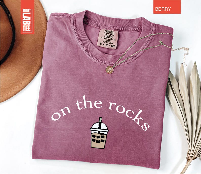 May include: Berry-coloured t-shirt featuring the words "on the rocks" and an iced beverage graphic. The shirt has a crew neck and short sleeves. A gold necklace with a pendant is draped across the shirt. The shirt is labelled "Comfort Colors".
