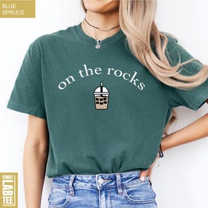 May include: Teal t-shirt featuring the words "on the rocks" in white, with a bubble tea graphic. The tee is a short-sleeved crew neck. The words "Blue Spruce" and "Street Lab Tee" are also visible on the shirt.