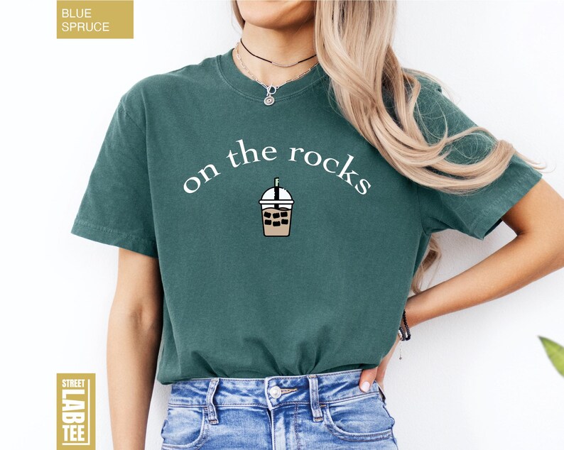 May include: Teal t-shirt featuring the words "on the rocks" in white, with a bubble tea graphic. The tee is a short-sleeved crew neck. The words "Blue Spruce" and "Street Lab Tee" are also visible on the shirt.