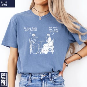 May include: Blue jean-colored t-shirt with white text and illustrations. The text reads "Do you have any fucks?" and "Not even one, girl. Go fish." The shirt features a graphic of two women seated at a table.