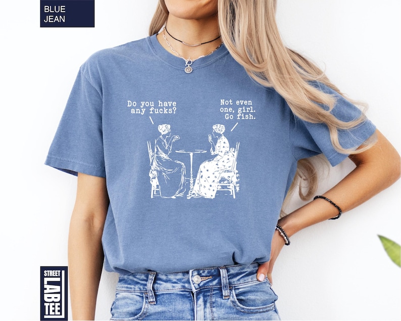 May include: Blue jean-colored t-shirt with white text and illustrations. The text reads "Do you have any fucks?" and "Not even one, girl. Go fish." The shirt features a graphic of two women seated at a table.