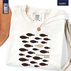 Puede incluir: Camiseta de color marfil con un dise&ntilde;o de peces. El dise&ntilde;o presenta un grupo de peces de color marr&oacute;n oscuro, con un pez en un tono m&aacute;s claro. El texto "CALLED TO BE DIFFERENT ROMANS 12:2" est&aacute; impreso debajo.