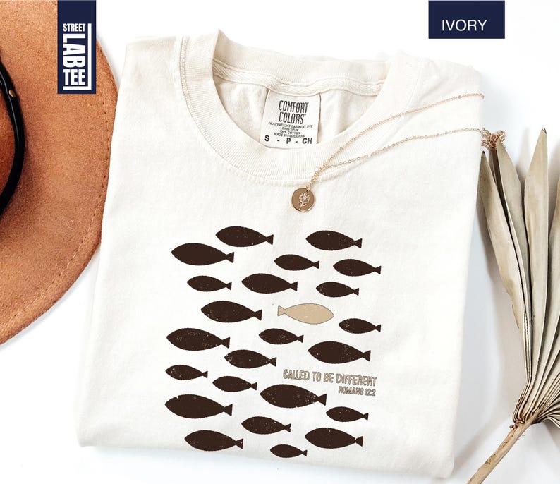 Puede incluir: Camiseta de color marfil con un dise&ntilde;o de peces. El dise&ntilde;o presenta un grupo de peces de color marr&oacute;n oscuro, con un pez en un tono m&aacute;s claro. El texto "CALLED TO BE DIFFERENT ROMANS 12:2" est&aacute; impreso debajo.