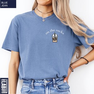 May include: A blue jean-colored t-shirt with the text "on the rocks" and a graphic of an iced coffee drink. The shirt is a short-sleeved, casual style.
