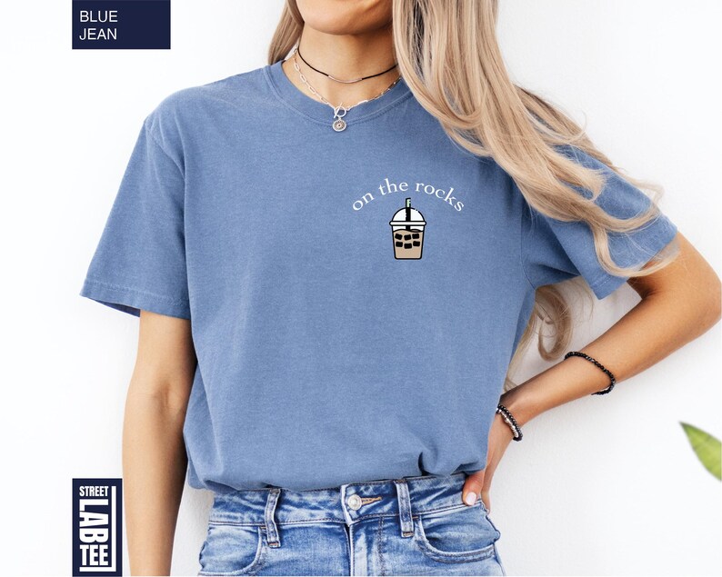 May include: A blue jean-colored t-shirt with the text "on the rocks" and a graphic of an iced coffee drink. The shirt is a short-sleeved, casual style.