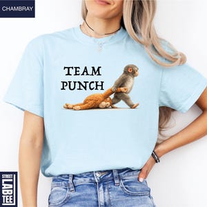 May include: Light blue t-shirt with the text "TEAM PUNCH" in black. The shirt features a cartoon illustration of two monkeys, one pulling the other. The shirt is made of a soft material.
