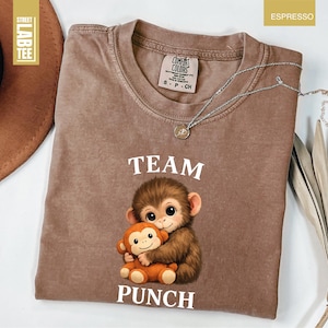 May include: Espresso-colored t-shirt featuring a graphic of two cartoon monkeys hugging, with the words "TEAM PUNCH" in white. The shirt has a crew neck and short sleeves. A silver necklace with a pendant is draped across the shirt.