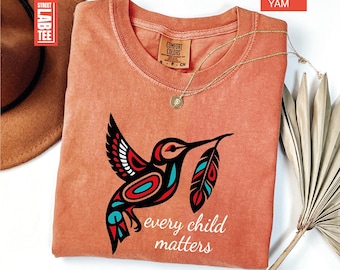 May include: A yam-coloured t-shirt with a crew neck and a hummingbird graphic. The hummingbird is black, red, and turquoise, holding a feather. The text "every child matters" is below the bird. A gold necklace is draped on the shirt.