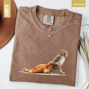 May include: Espresso-colored t-shirt with a crew neck and a graphic of two monkeys. One monkey is pulling the other. The shirt has a Comfort Colors tag. A silver necklace with a pendant is on the shirt.