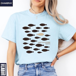 Puede incluir: Camiseta azul claro con un gr&aacute;fico de peces, uno de ellos de diferente color. El texto "CALLED TO BE DIFFERENT ROMANS 12:2" est&aacute; impreso debajo del gr&aacute;fico de peces. La camiseta es de un material suave.