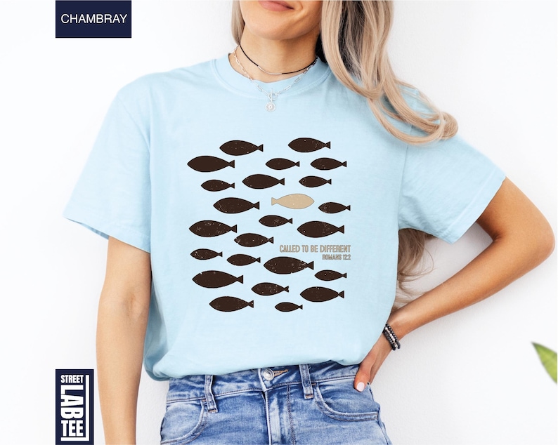 Puede incluir: Camiseta azul claro con un gr&aacute;fico de peces, uno de ellos de diferente color. El texto "CALLED TO BE DIFFERENT ROMANS 12:2" est&aacute; impreso debajo del gr&aacute;fico de peces. La camiseta es de un material suave.