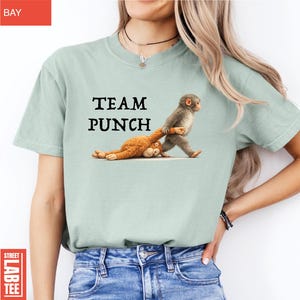 May include: A light green t-shirt with the text "TEAM PUNCH" above a cartoon image of two monkeys. One monkey is pulling the other by the arm. The shirt is a solid color.